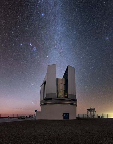 The soft glow of the Milky Way pours down like a waterfall over the VLT Survey Telescope (VST) at ESO’s Paranal Observatory. 2635 metres above sea level, the VST has an unparallelled view of the magnificently clear skies above Chile’s Atacama Desert. It is the largest telescope in the world dedicated to observational surveys in visible light, and it contributes to a vast range of studies, from discovering remote Solar System bodies to searching for exoplanet transits, to studying the structure and evolution of our galaxy. Above the VST, a few stars are particularly prominent just outside the main band of the galaxy: the bright reddish star is Betelgeuse, a red supergiant 640 light-years away thought to be on the brink of a supernova explosion. Betelgeuse and Bellatrix, the fainter white star to its left, are the shoulders of Orion the Hunter, one of the most famous and easily recognisable constellations in the sky. The three stars forming a straight line above the shoulders are Orion’s belt, and the nearby purple-tinged cluster of light is the Orion Nebula. Though it looks like a fuzzy patch to the naked-eye, binoculars or a telescope reveal it to be a spectacular nebula, host to massive amounts of star formation.
